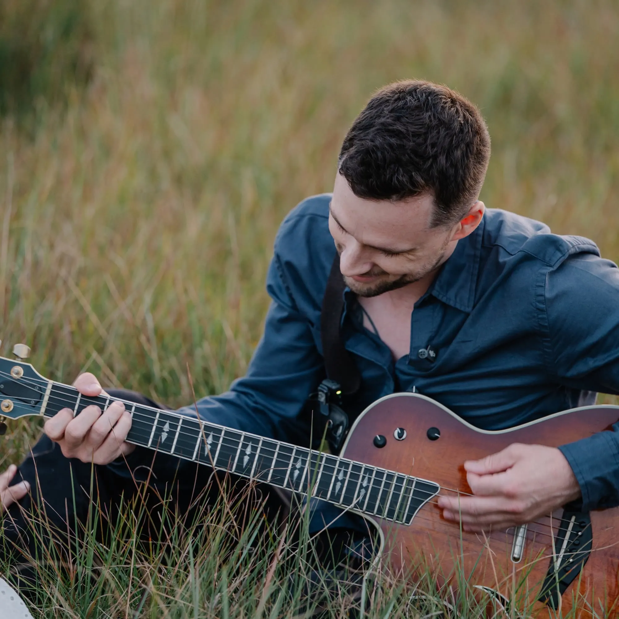 Noah Spott sitting in a field of tall grass playing a sunburst acoustic-electric guitar